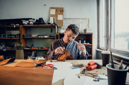 Sitting at a table in the workshop a young guy manually sews leather elements to each other.の写真素材