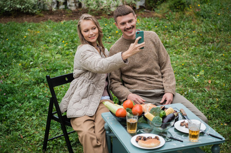 A guy and a girl take a selfie together on a smartphone while sitting at a picnic table. On the lawn near the house on a warm autumn dayの写真素材