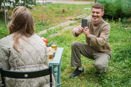 The husband takes photos of his wife on a smartphone. A woman is sitting at a picnic table, which is...