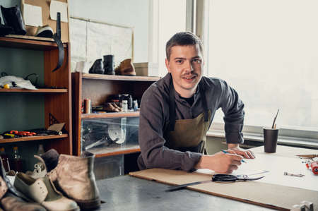 A young shoemaker makes a drawing for a pattern for leather shoes on a table in his workshop.の写真素材