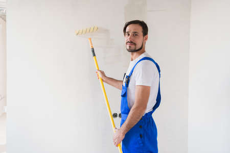 A bearded builder in work clothes paints the wall with a roller. He smiles friendly and poses for the cameraの写真素材