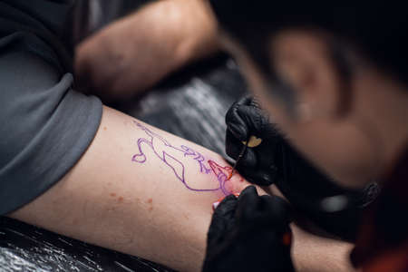 Close-up photo of a girl tattoo artist putting a tattoo on the arm, of a young guy in a tattoo...