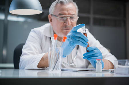 Office of the researcher of antiviral drugs. An elderly chemist doctor holds a test tube with a medical preparation in his hands.の写真素材