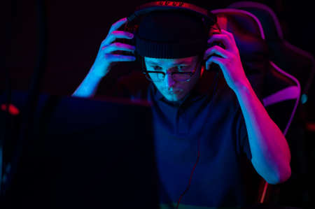 A young esports player sits at a computer during an online game tournament. Neon lighting of the esports arena. A hat and headphones with a headset.の写真素材