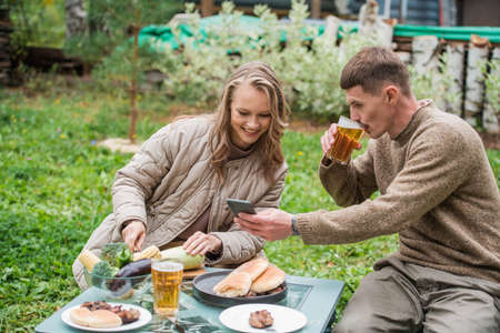 A close-knit family, husband and wife, together watch video content on a smartphone during a picnic in nature near a country house. Grilled meat, vegetables, and beer on the table.の写真素材