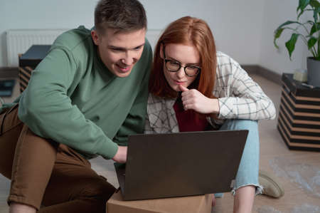 Newlyweds pick up furniture for their new apartment, sit on the floor with a laptop among cardboard boxesの写真素材