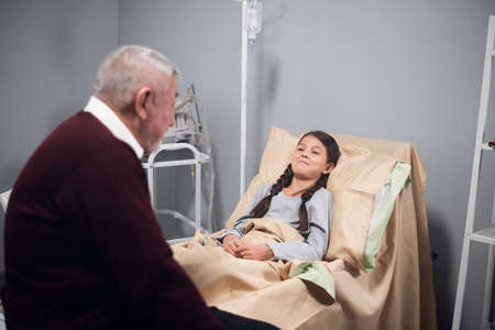 medicine, support, family health care and people concept. Happy senior man visiting his granddaughter, who is lying in bed at a hospital wardの写真素材