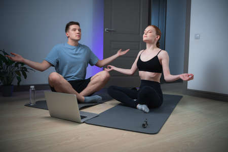 During an online yoga class, a young couple meditates on gym mats at home in front of a laptopの写真素材