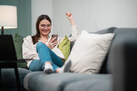 A pretty freelancer girl with glasses lies down on the sofa cushions, holds a phone in her hands and communicates with friendsの写真素材