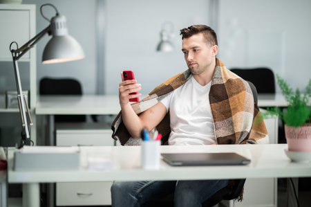 In the office, the manager falls ill, covers himself with a blanket and calls the doctor to report the symptoms of the diseaseの写真素材