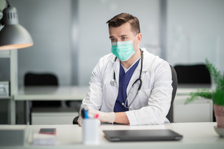 During the covid-19 pandemic, a doctor sees patients in his office wearing a medical maskの写真素材