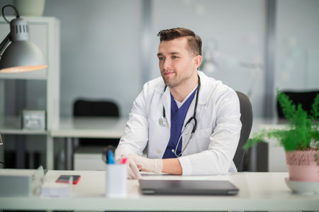 A cute young doctor is sitting at his desk with a stethoscope and gloves onの写真素材