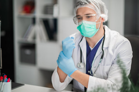 A young male doctor wearing a mask, medical cap and glasses puts on blue gloves before receiving a doctor in his officeの写真素材