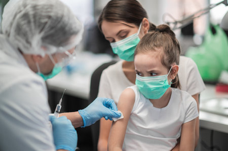 A little girl is afraid of the sight of a needle and pain during vaccination in a medical laboratory, her mother supports herの写真素材