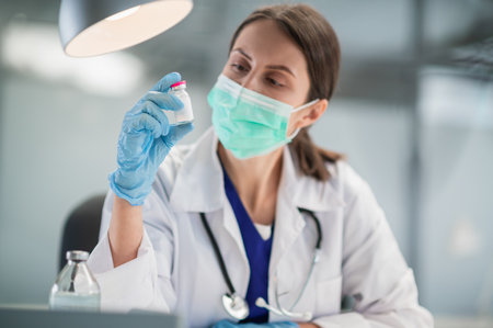 A young woman doctor in a medical mask in the clinic holds a bottle of coronavirus vaccine in her handsの写真素材