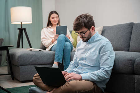 A young couple of freelancers working from home on the Internet, a girl on the couch holding a tablet, a guy on the floor working on a laptopの写真素材