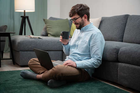 During the isolation in the coronavirus quarantine, the guy sits on the floor, works remotely in a laptop and drinks from a coffee mugの写真素材