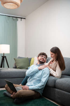 A young girl helps her bearded, bespectacled boyfriend working from home who is sitting on the floorの写真素材