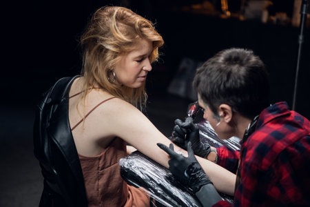 A blonde woman in a tattoo parlor makes a tattoo on her arm in the form of a word, tattooist young girlの写真素材