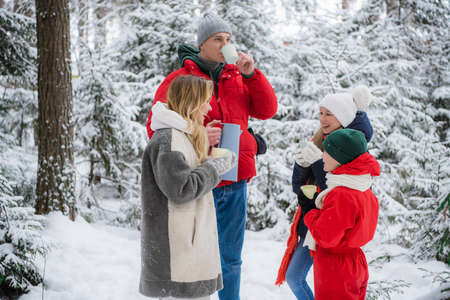 A healthy friendly family actively spends a weekend in nature in a winter spruce forest during a beautiful snowfall and enjoys hot tea from hiking mugs.の写真素材