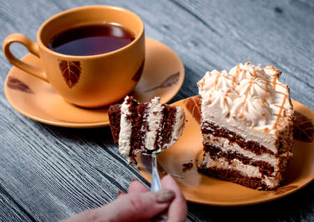On grey wooden table is a plate with a slice of cake and a Cup of tea. Visible hand with a teaspoon. On the spoon is the piece of cake.の写真素材