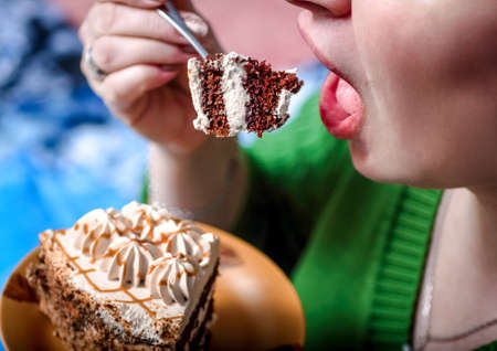 The woman opens her mouth to eat a piece of cake. In her hand she holds a plate with a cake