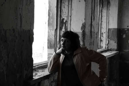 A young girl stands by the window in a ruined old house. Black and white photo.の写真素材