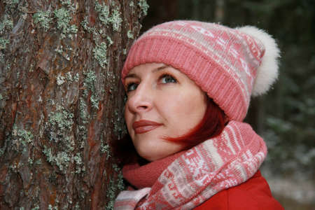 A young woman in a hat and scarf stands near a tree in the winter forest.