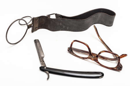 Old and worn rusty razor, sharpening leather, glasses on a white background