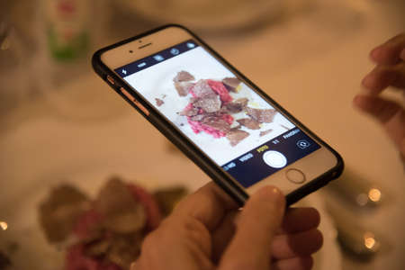 Close-up view of woman hand with smartphone photographing plate with meat and truffle to place...