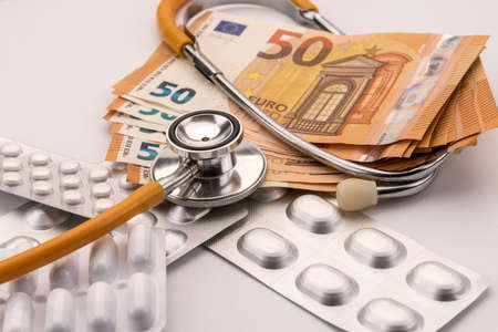 euro banknotes and blister packs of medicines in capsules and pills with stethoscope above, on white. Health care cost conceptの写真素材