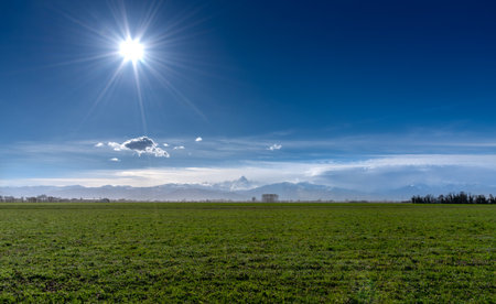Landscape of green meadows in the upper Po Valley, Piedmont, Italy, with Mount Monviso in the clouds in the background with blue sky and sunshineの写真素材