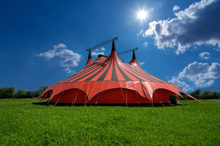 Red and black circus tent in green meadow with blue sky with clouds and sunshineの写真素材
