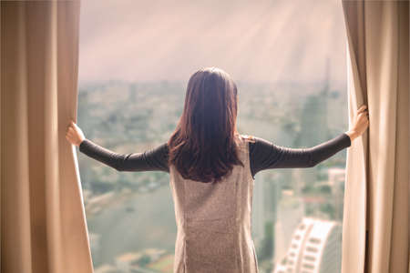 Asian portrait beautiful woman opening curtains and building cityscape backgroundの写真素材