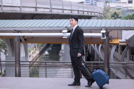 Asian Businessman smiling and happy, wearing suit with luggage, walking on business street for travelling on vacation tourist business trip conceptの写真素材