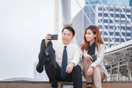 businessman sitting and using phone to take photo as selfie with colleague together and modern office building backgroundの写真素材