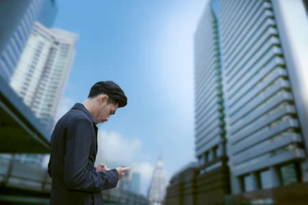 asian business man standing and use smartphone with business building background