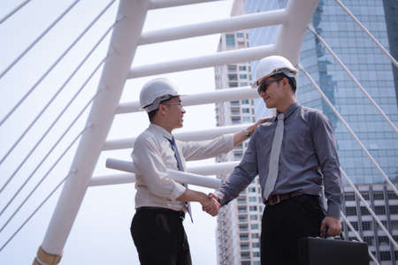 Asian businessman discuss and shake hand with engineer architect professional wearing safety helmet and suite, holding construction roll plan and briefcase for working team conceptの写真素材