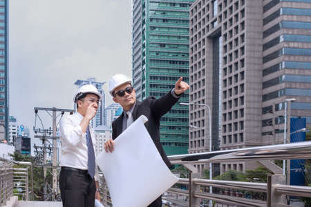 Asian businessman looking and point to finger away and engineer architect hold construction industrial plan background for working together as team conceptの写真素材