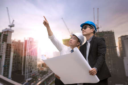 Asian businessman looking and point to finger away and engineer architect hold construction industrial plan background for working together as team conceptの写真素材