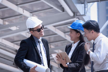 Asian businessman and woman discuss with engineer architect professional wearing safety helmet and suite, looking at tablet and holding construction roll plan for working team conceptの写真素材