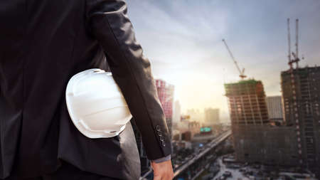 White helmet of engineer architect holding by businessman in suit and building construction site backgroundの写真素材