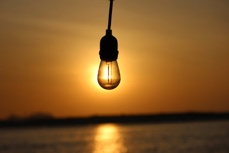 A vintage-style incandescent light bulb is silhouetted against a vibrant golden sunset sky, with the...