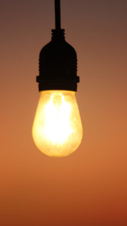 A close-up vertical shot of a single incandescent light bulb with a glowing filament, suspended...