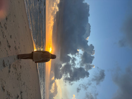 Rear view of a person wearing a hijab and modest clothing, walking along a sandy beach towards the rising sun. The sun is low on the horizon, casting a golden glow over the ocean and creating reflections on the wet sand. Dramatic clouds are visible in theのeditorial素材