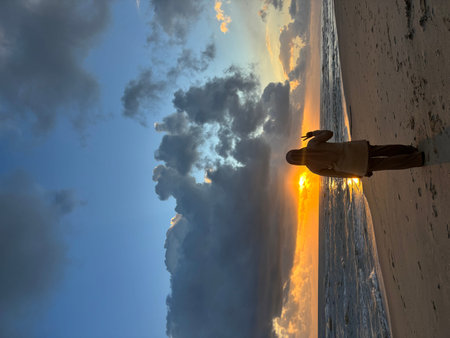 A woman wearing a hijab stands on a sandy beach, seen from behind, facing the ocean during sunset. She holds up two fingers in a peace sign gesture. The sun sets over the sea, casting a golden glow on the water and horizon, with dramatic clouds filling thのeditorial素材