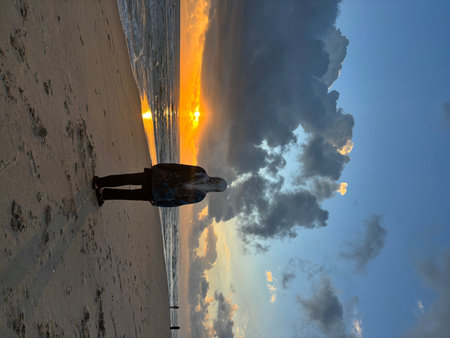 A person wearing a hijab stands on a sandy beach, gazing at the vibrant sunrise over the ocean. The golden sun reflects brilliantly on the water, illuminating the waves and scattered clouds in the sky. Other figures are visible in the distance along the sのeditorial素材