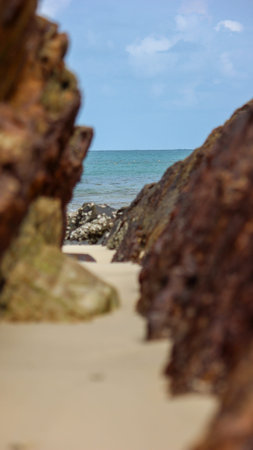 A serene view of the sea from a sandy beach, with large, out-of-focus brown rock formations framing the turquoise water and a clear blue sky. Small buoys dot the distant horizon.のeditorial素材