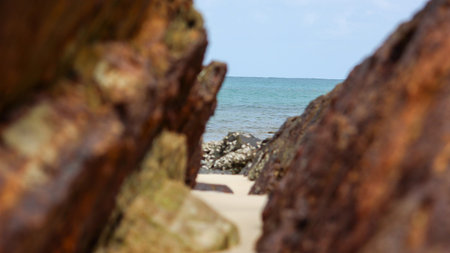 A scenic view of the calm, turquoise sea and sandy beach, framed by large, out-of-focus rusty rocks. Smaller rocks covered in barnacles are visible on the beach. The horizon shows a clear sky and distant ocean.のeditorial素材