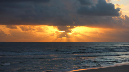View of a dramatic sunset or sunrise over the ocean, featuring golden light breaking through dark, heavy clouds. Sunrays illuminate the water and sky, with gentle waves rolling onto a sandy beach in the foreground.のeditorial素材